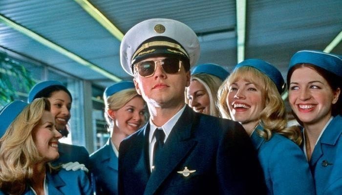 Still image from the film "Catch Me If You Can" showing Leonardo DiCaprio's character, Frank Abignale, posing as an airline pilot with a group of smiling air hostesses